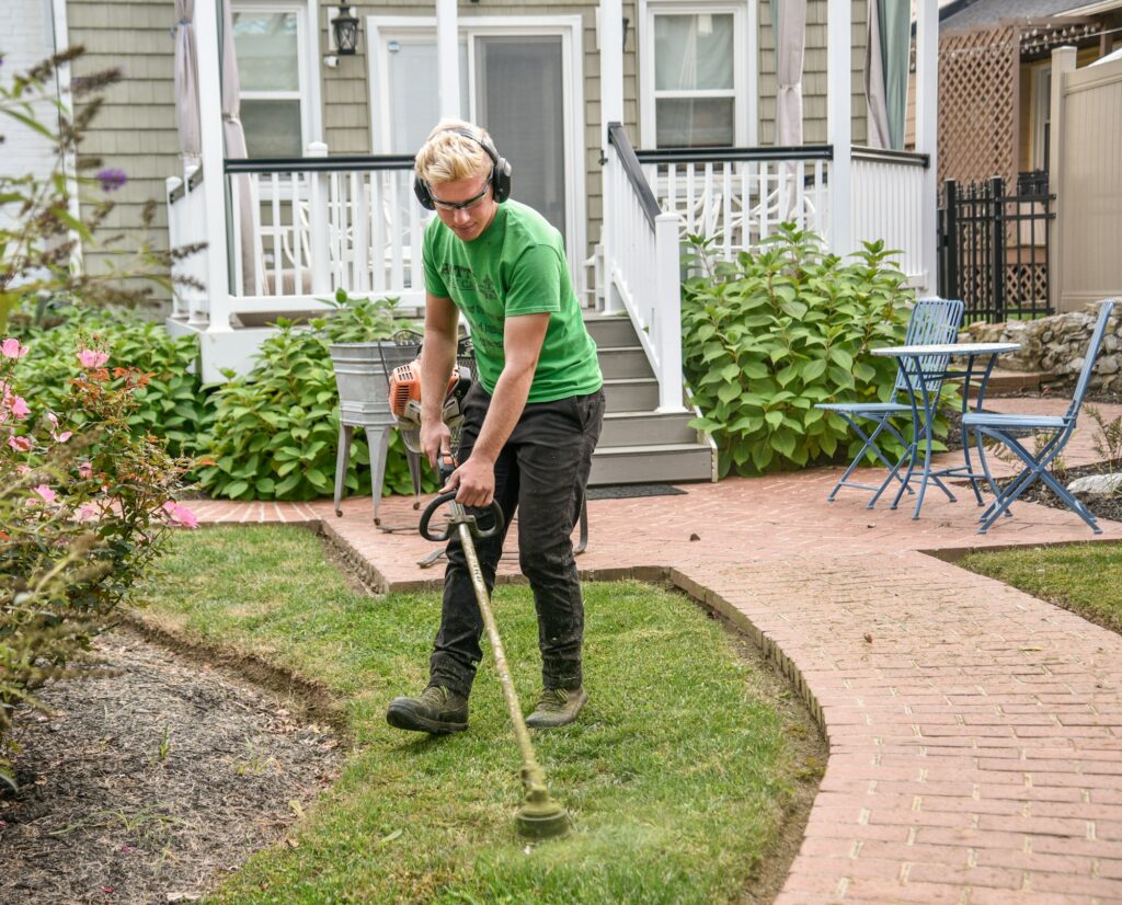 A man in ear protection running a string trimmer along a lawn edge in a front yard beside a brick walk