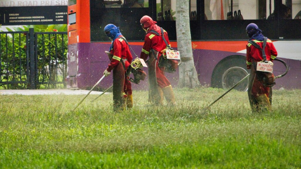 Three grounds workers in face shields, gloves and boots running string trimmers along a roadside lawn